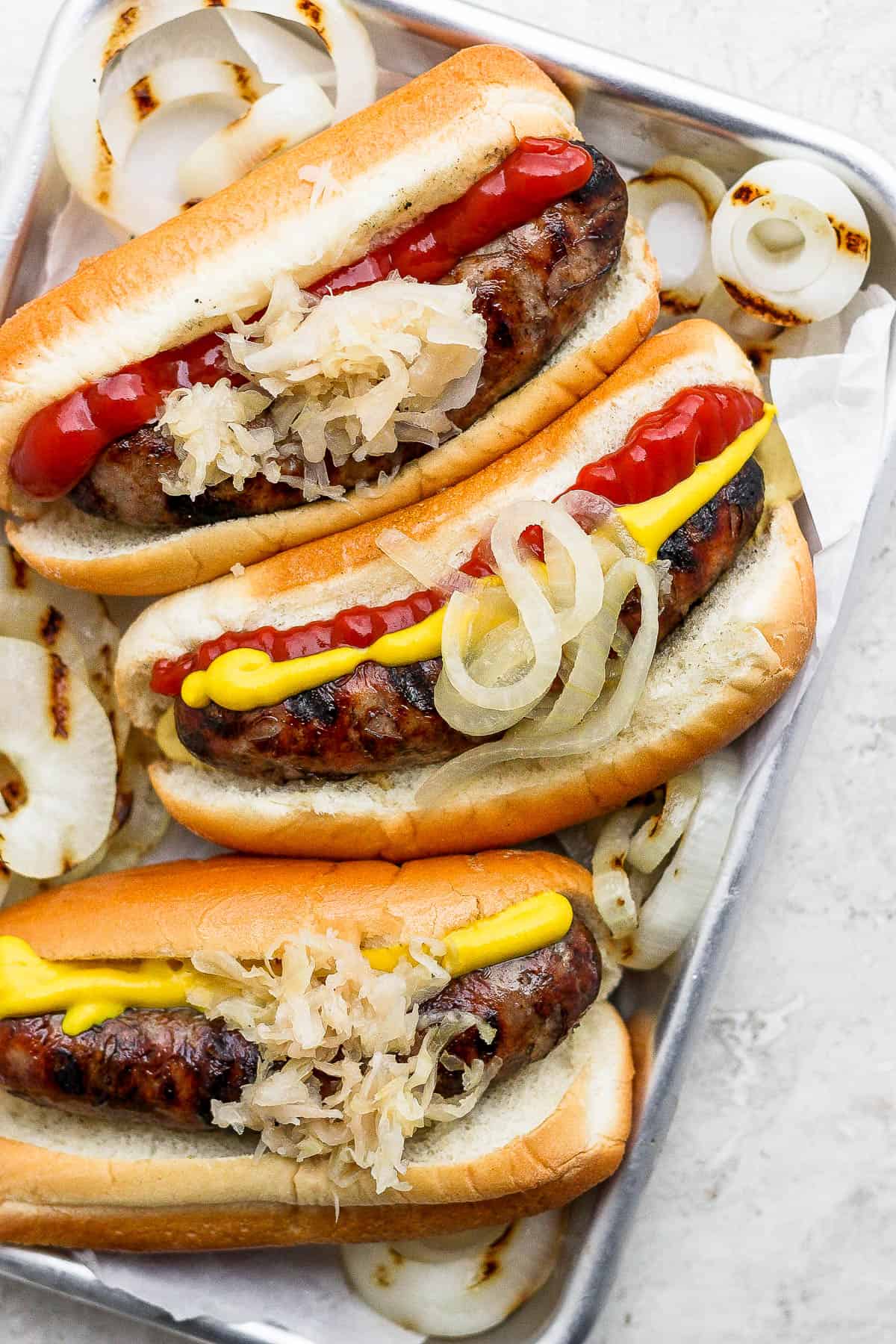 Grilled brats in buns on a metal tray with some grilled onions.