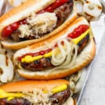 Top down shot of a small pan of 3 grilled brats in buns with onion, ketchup and mustard.