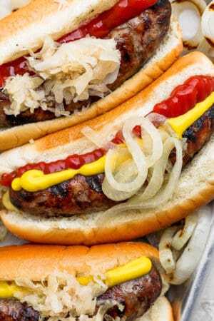 Top down shot of a small pan of 3 grilled brats in buns with onion, ketchup and mustard.