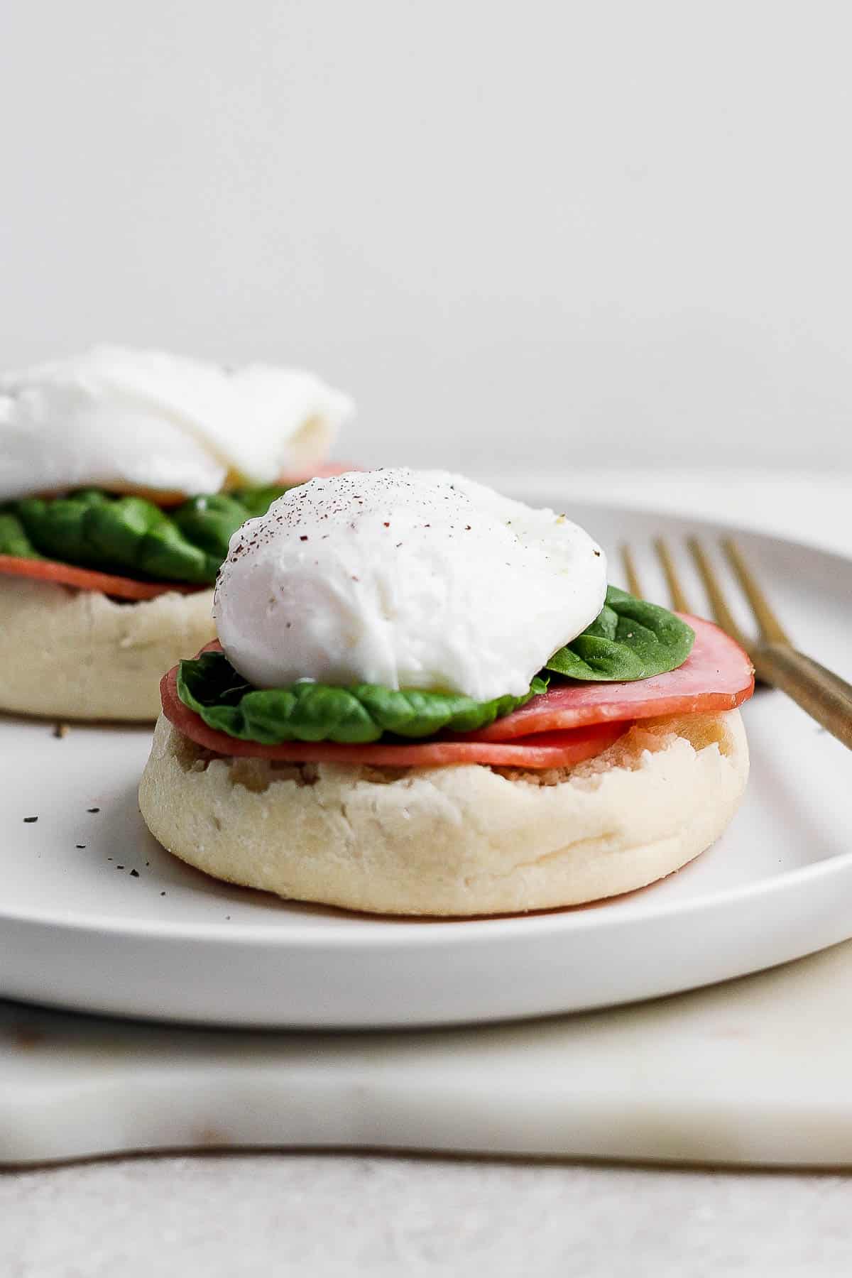 A white plate with english muffin halves topped with Canadian bacon, fresh spinach, and poached egg.