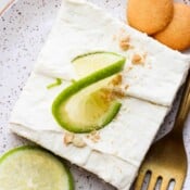 Top down shot of a key lime cheesecake bar on a speckled plate with a lime on top and next to it and two vanilla wafers on the other side.