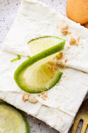 Top down shot of a key lime cheesecake bar on a speckled plate with a lime on top and next to it and two vanilla wafers on the other side.