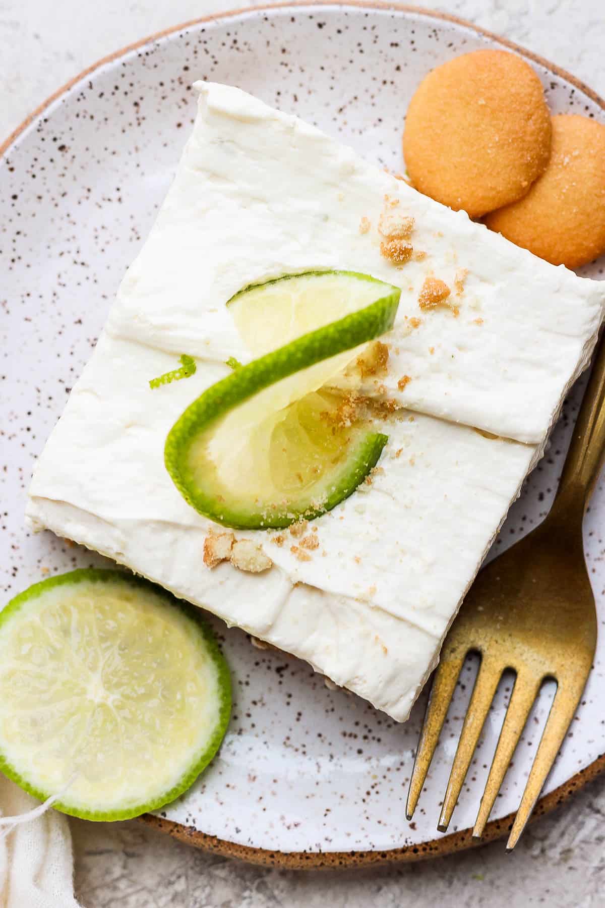 A key lime cheesecake bar on a plate topped with a slice of lime and crumbled vanilla wafers.