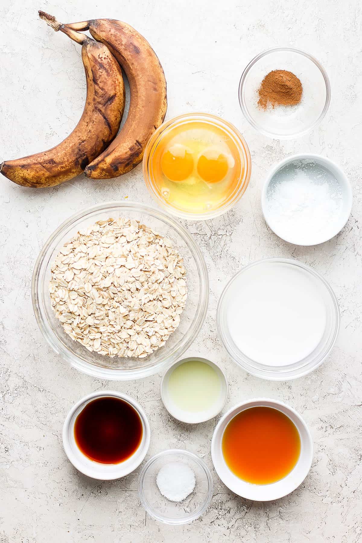 An ingredient shot showing two ripe bananas and individual containers of egg, oats, cinnamon, almond milk, vanilla, salt, baking powder, and mini chocolate chips.
