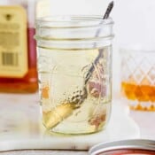 A mason jar filled with homemade simple syrup with a spoon sticking out and a bottle of whiskey in the background.