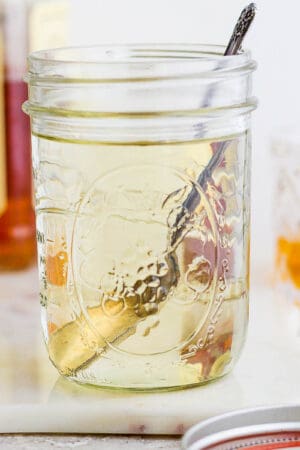 A mason jar filled with homemade simple syrup with a spoon sticking out and a bottle of whiskey in the background.
