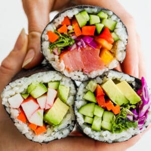Two hands holding together 3 different types of sushi burritos: spicy tuna, california and avocado.