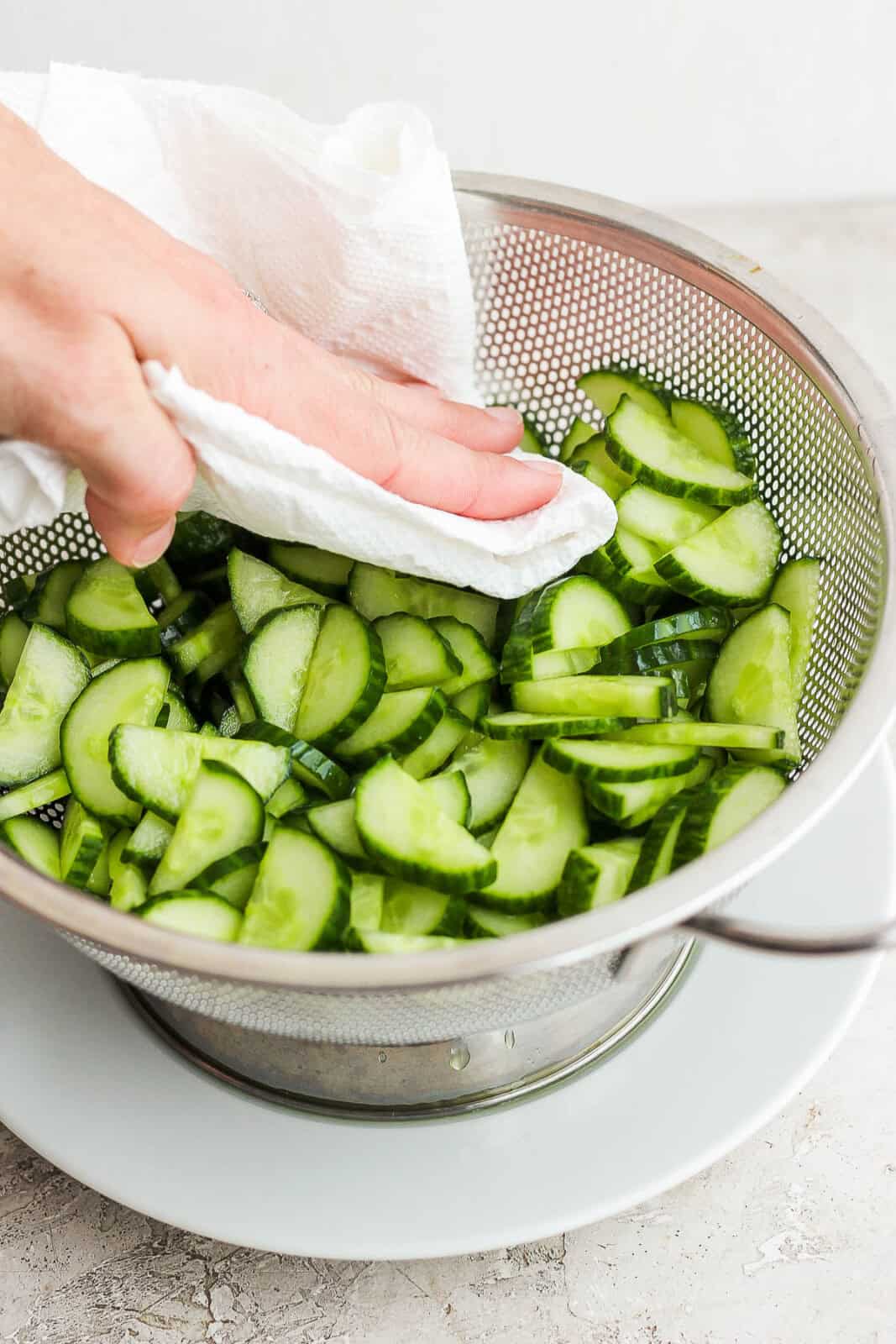 A hand patting sliced and halved cucumbers dry with a paper towel to make creamy cucumber salad with sour cream and mayo.