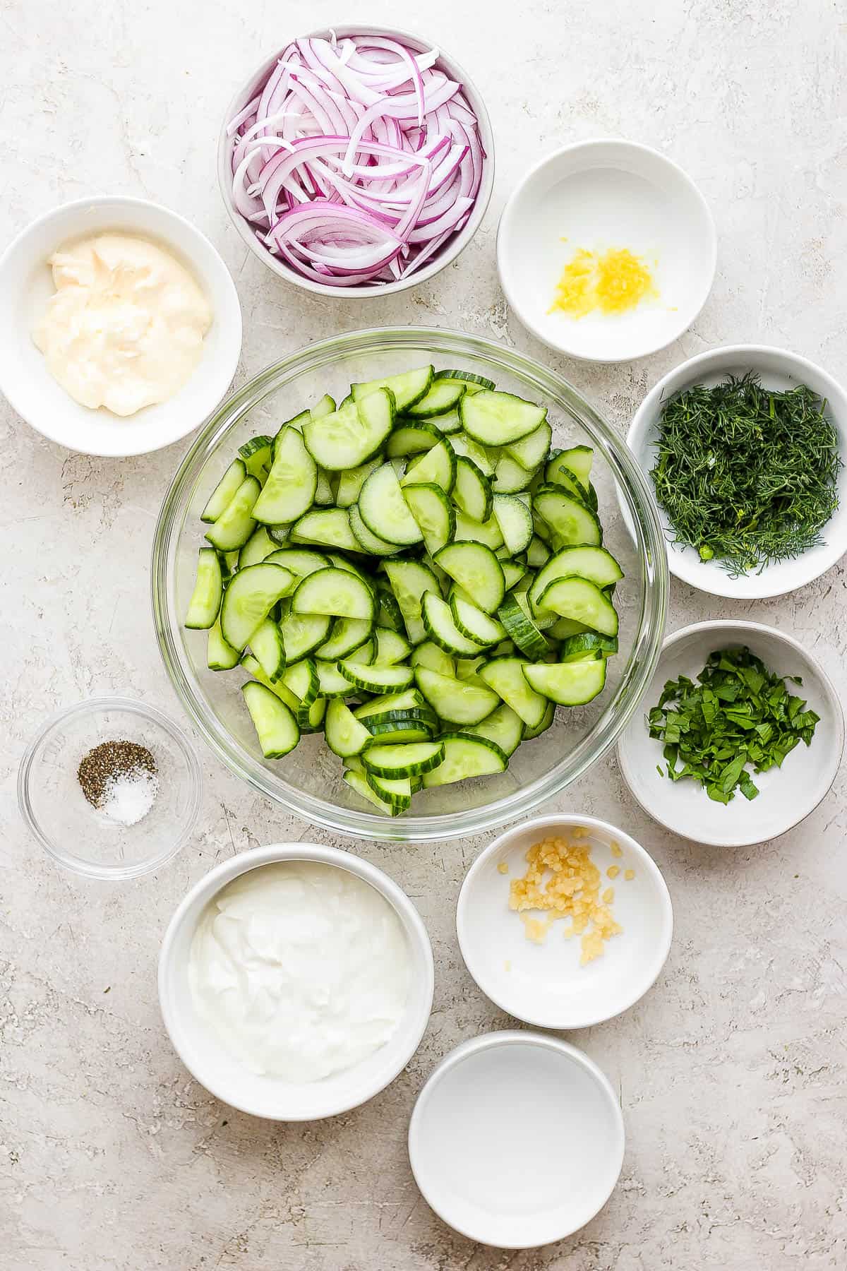Creamy cucumber salad ingredients, each in their own bowl; mayonnaise, red onion, garlic, dill, sour cream, white vinegar, parsley, lemon juice, lemon zest, ground black pepper, and salt.