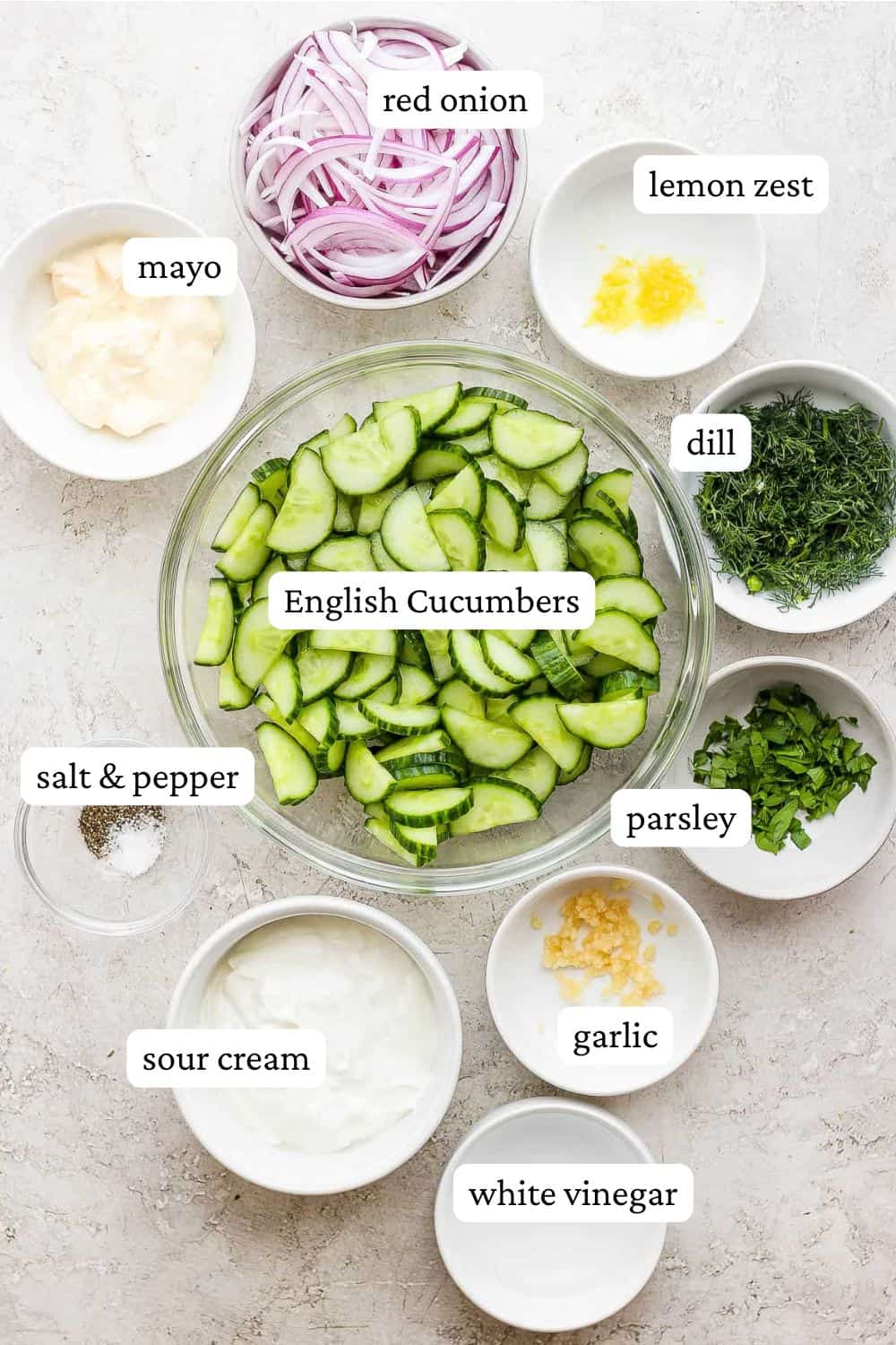 creamy cucumber salad with sour cream and mayo ingredients in individual bowls including red onion, lemon zest, mayo, sliced and halved cucumbers, fresh dill, fresh parsley, salt and pepper, minced garlic, sour cream, and white vinegar.