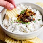 A bowl of homemade french onion dip with caramelized onions on top and someone using a chip to scoop up some dip.
