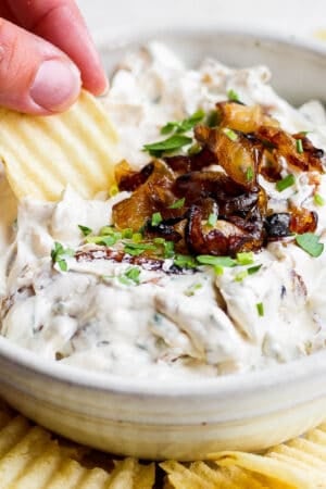 A bowl of homemade french onion dip with caramelized onions on top and someone using a chip to scoop up some dip.