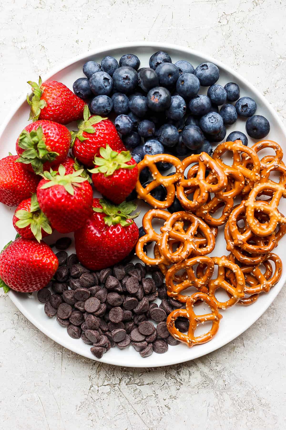 A plate full of strawberries, chocolate chips, pretzels, and blueberries.
