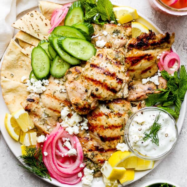 A plate of grilled greek yogurt chicken with pickled onion, lemon, cucumber, pita bread, fresh herbs and tzatziki sauce.