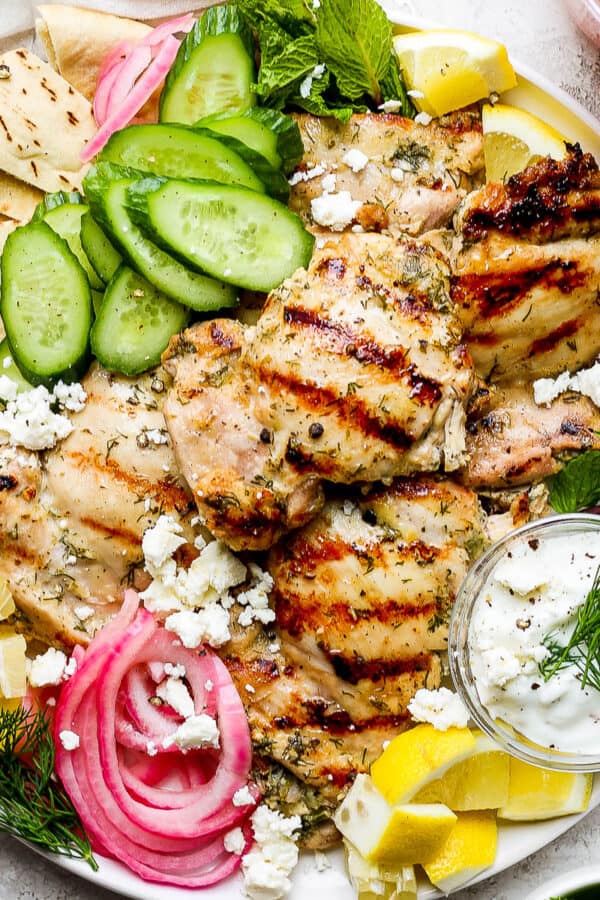A plate of grilled greek yogurt chicken with pickled onion, lemon, cucumber, pita bread, fresh herbs and tzatziki sauce.