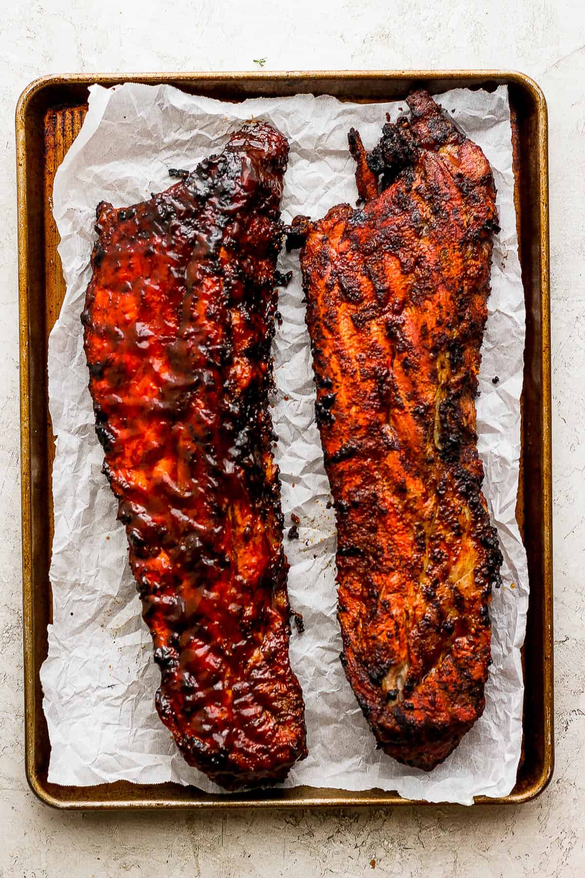 Grilled ribs resting on a baking sheet, one has bbq sauce on it and one does not.