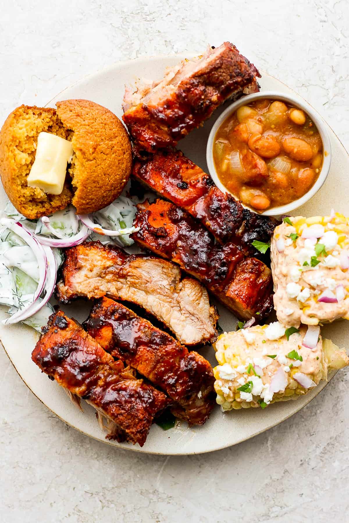 A plate full of grilled ribs, slow cooker baked beans, creamy cucumber salad, Mexican street corn salad, and a cornbread muffin.