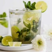 Straight on shot of a glass of mojito on a white plate with a flower next to it. The mojito has mint sticking out of it and a lime on the rim.