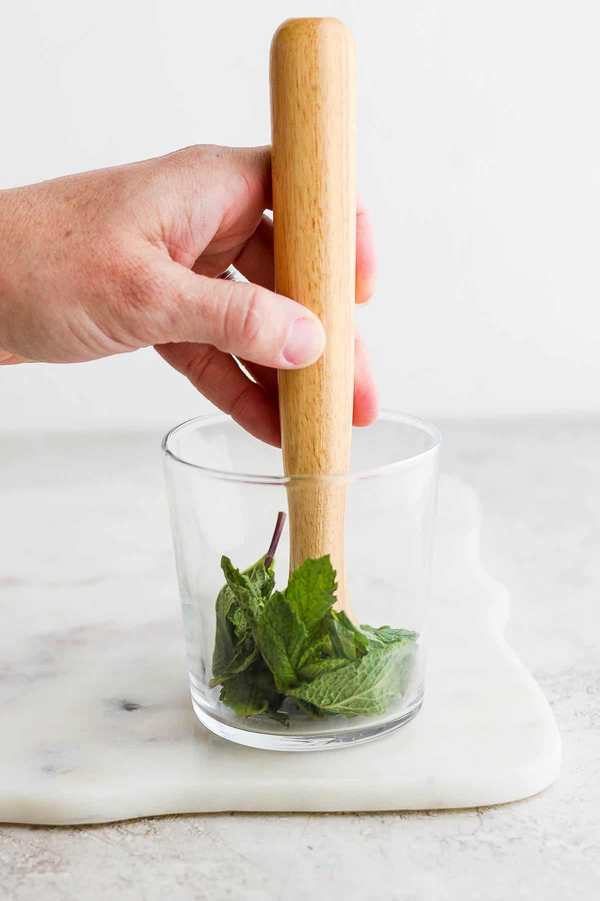 Mint leaves being muddled in the bottom of a clear glass.