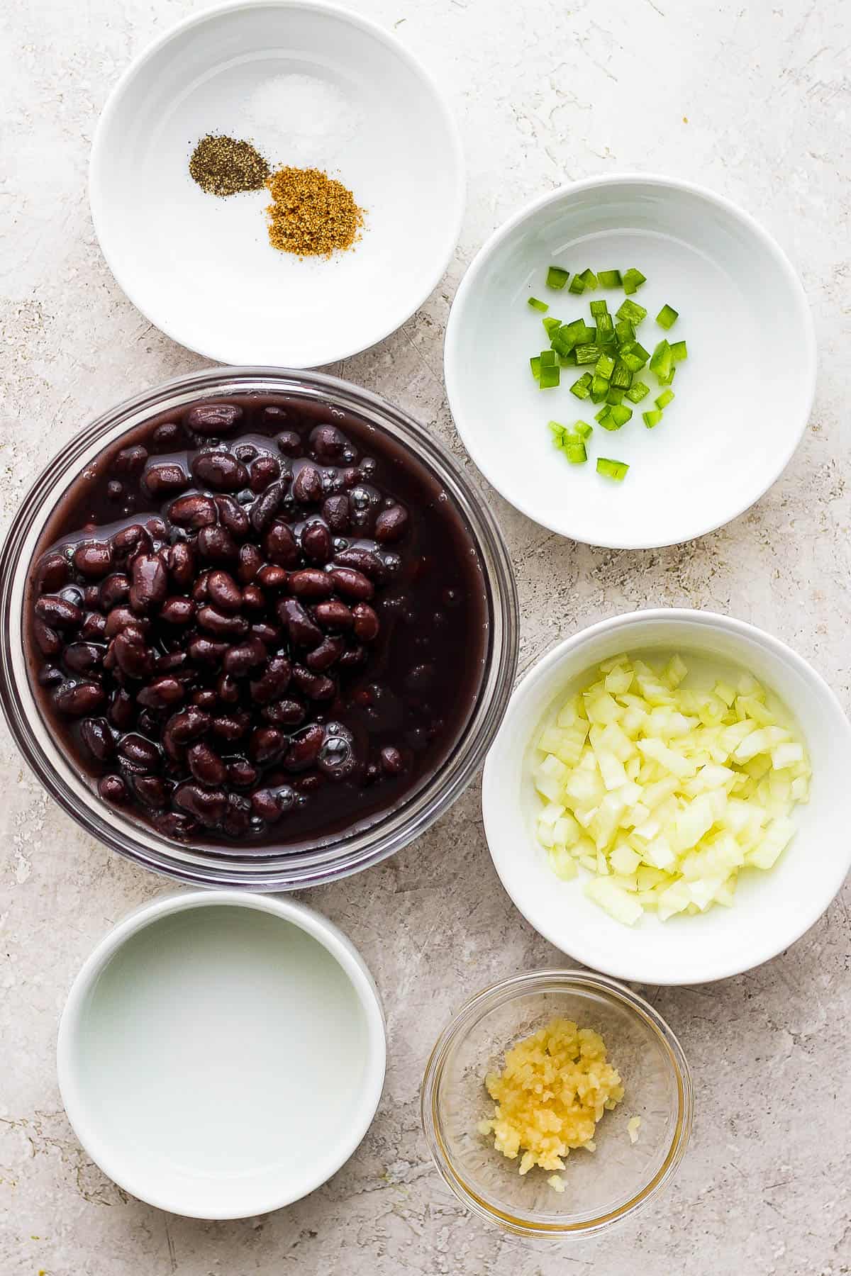 Individual ingredient bowls of black beans, cumin, salt, pepper, jalapeno, onion, garlic, and olive oil.