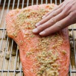 Side angle shot of a someone rubbing a smoked salmon brine on top of a salmon fillet placed on a wire rack on top of a baking sheet.