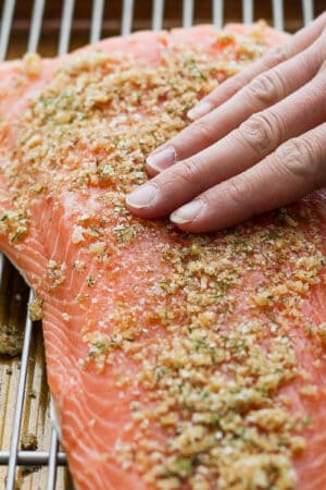 Side angle shot of a someone rubbing a smoked salmon brine on top of a salmon fillet placed on a wire rack on top of a baking sheet.