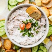 Top down shot of a bowl of smoked salmon dip with chunks of salmon and chives in the middle. A few crostini pieces of bread sticking out and cucumber and lemons surrounding the bowl.