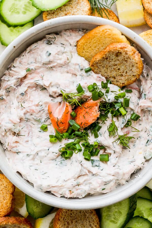 A bowl of smoked salmon dip with chunks of salmon and chives in the middle. A few crostini pieces of bread sticking out and cucumber and lemons surrounding the bowl.