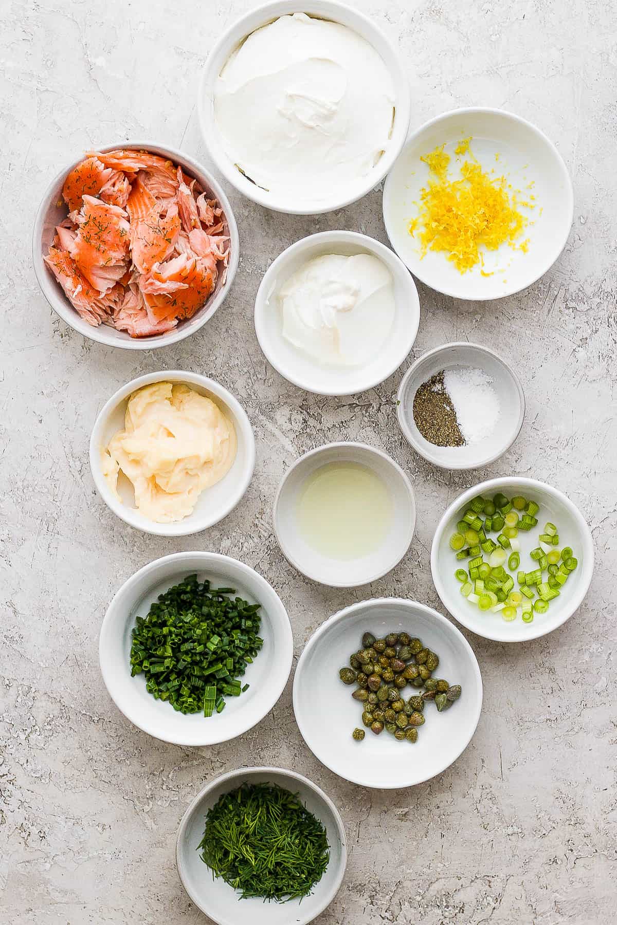 Eleven ingredient bowls each with a separate ingredient: smoked salmon, lemon zest, lemon juice, capers, chives, scallions, mayo, sour cream, dill, salt and pepper.