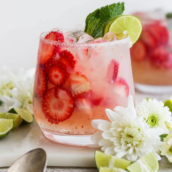 A strawberry mojito in a glass with extra strawberry slices and mint and lime garnish with flowers around the glass.