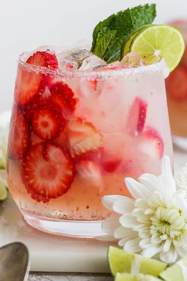 A strawberry mojito in a glass with extra strawberry slices and mint and lime garnish with flowers around the glass.