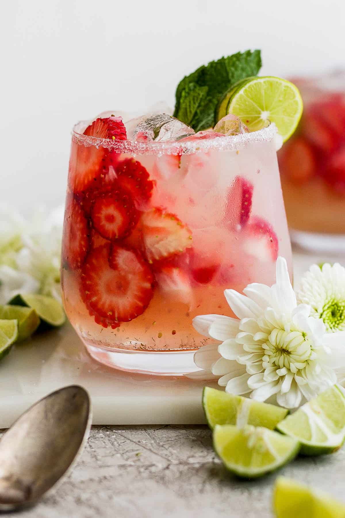 A strawberry mojito in a class surrounded by flowers and lime wedges.