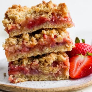 A stack of three strawberry rhubarb bars on a plate with two fresh strawberries next to them.