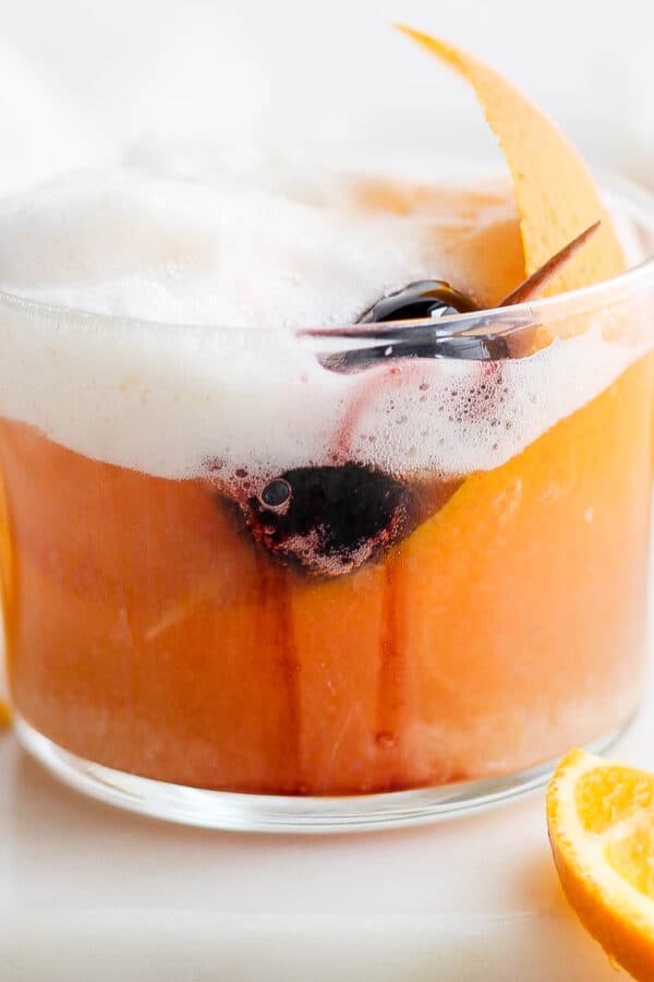 A short glass of cranberry orange whiskey sour with cocktail cherries and orange garnish.