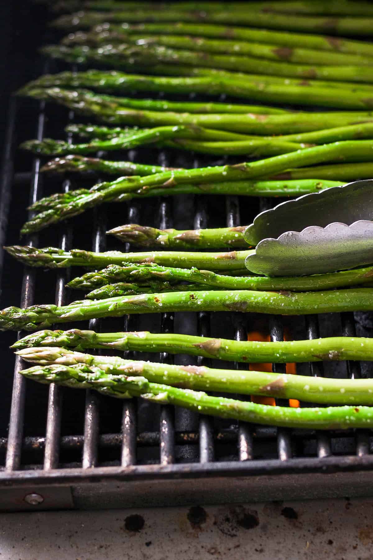 Asparagus cooking on the grill grates and a pair of tongs rotating them.