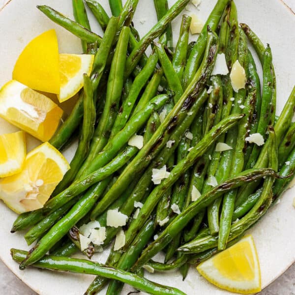 Plate of grilled green beans with lemon wedges and flakes parmesan cheese.