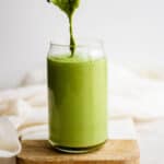 A glass with matcha smoothie in it and more being poured into it while it sits on a wooden board.