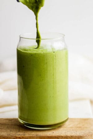 A glass with matcha smoothie in it and more being poured into it while it sits on a wooden board.