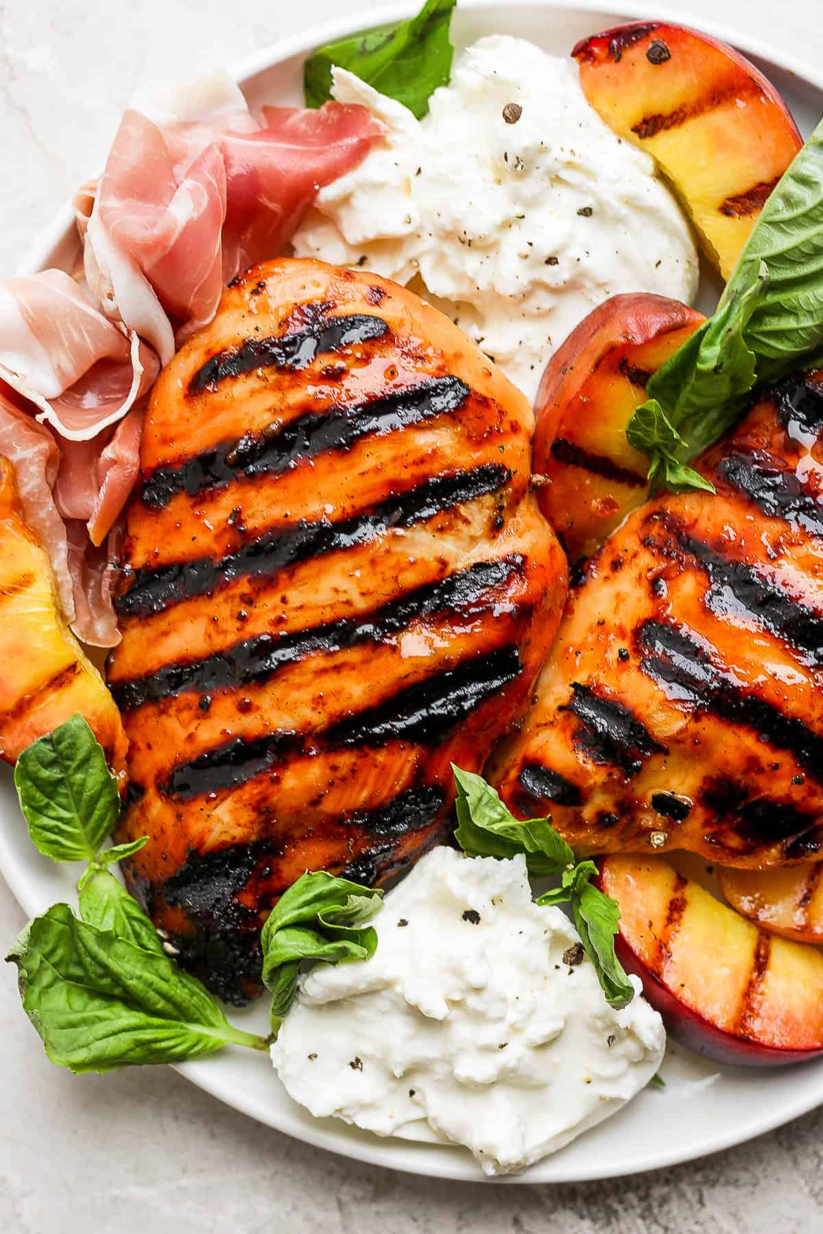Two grilled chicken breasts on a plate garnished with fresh basil leaves, burrata cheese, grilled peaches, and prosciutto.