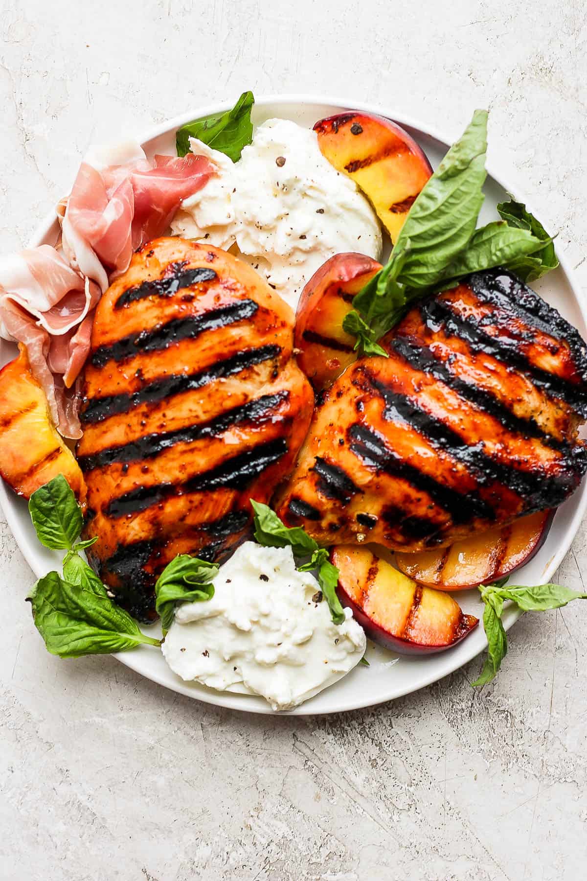 Two grilled chicken breasts surrounded by grilled peaches, burrata cheese, prosciutto, and fresh basil leaves.