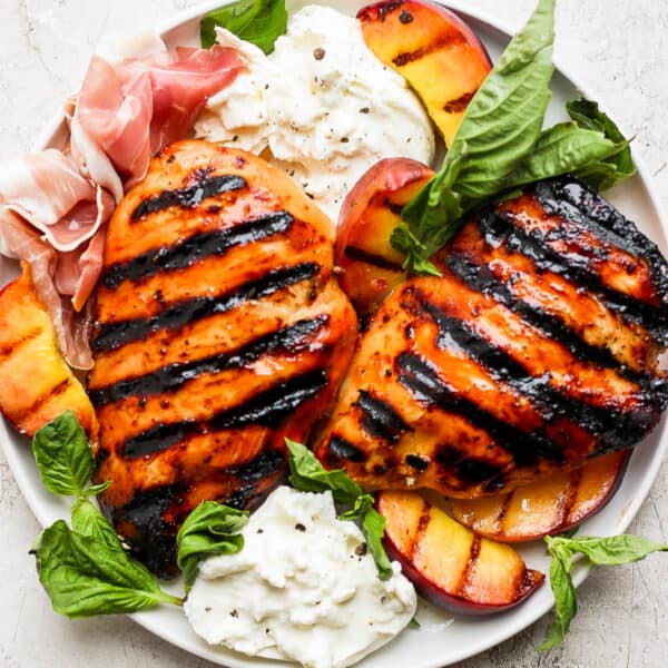 Grilled peach chicken on a plate with fresh basil, grilled peaches, fresh prosciutto and burrata cheese.