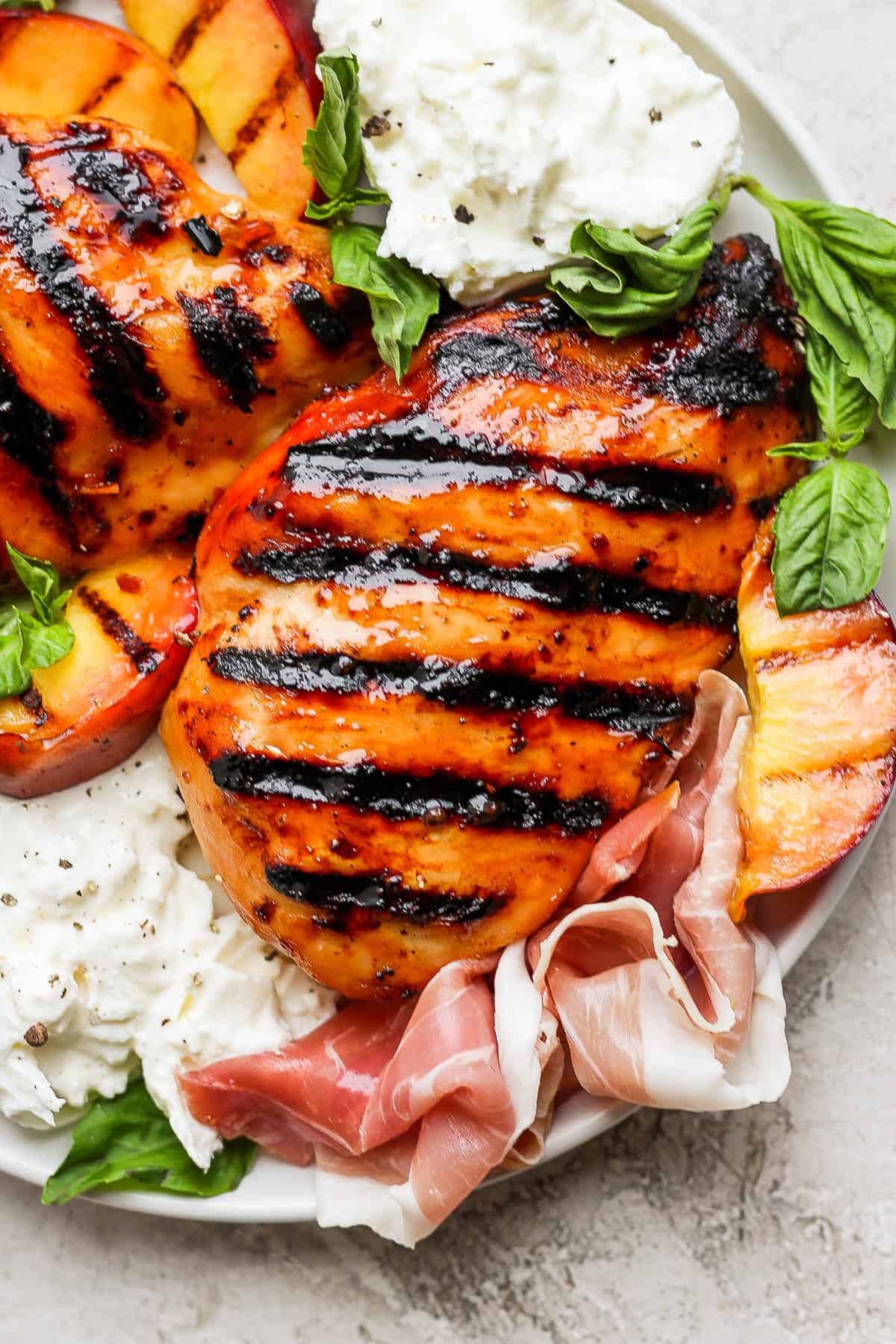 One grilled chicken breast surrounded by grilled peaches, burrata cheese, prosciutto, and fresh basil leaves.