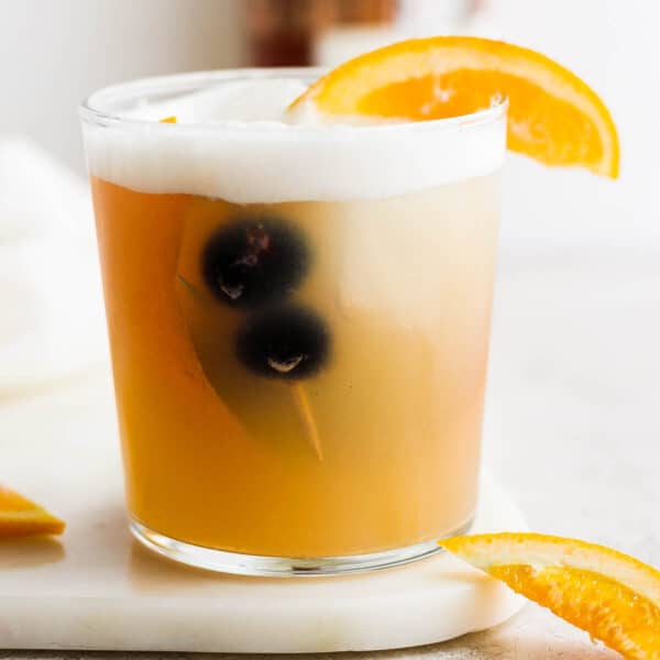 A glass on a marble board filled with a whiskey sour with two cocktail cranberries inside and an orange garnish.