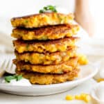 A stack of corn fritters on a plate with fork next to it and dollop of sour cream on top with minced chives.