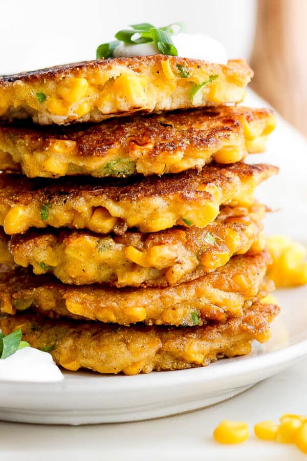 A stack of corn fritters on a plate with fork next to it and dollop of sour cream on top with minced chives.