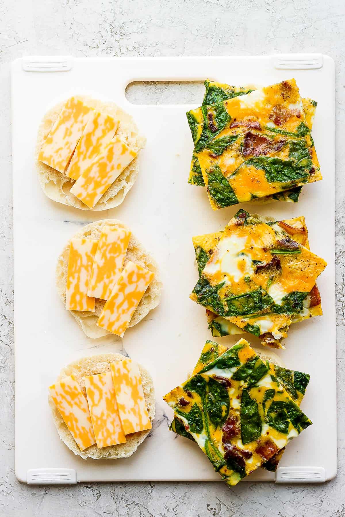 One side of a cutting board has the english muffin tops with cheese and the other side has the english muffin bottoms with egg squares on top.