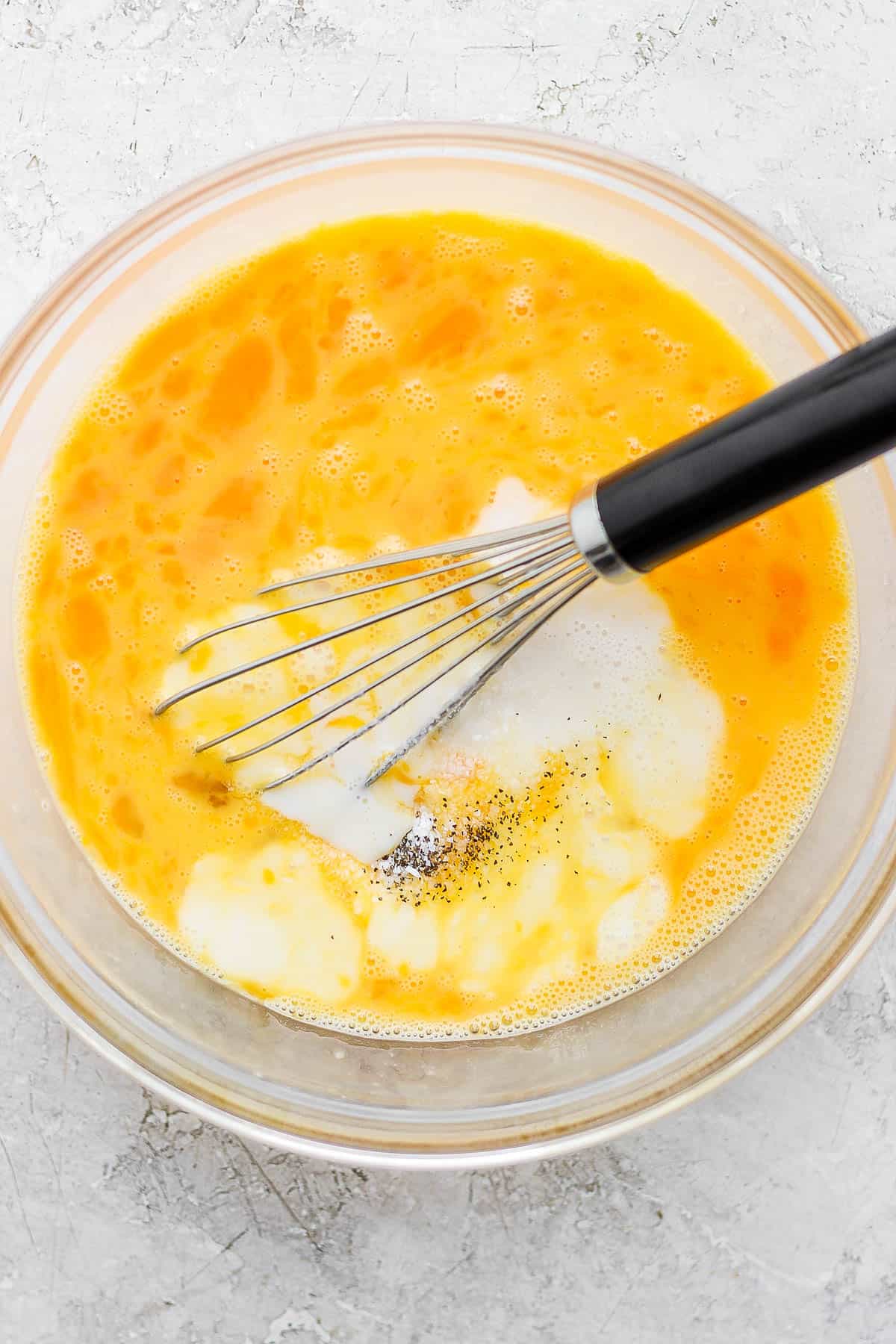 A large glass bowl with eggs, milk, salt, and pepper in it with a whisk.