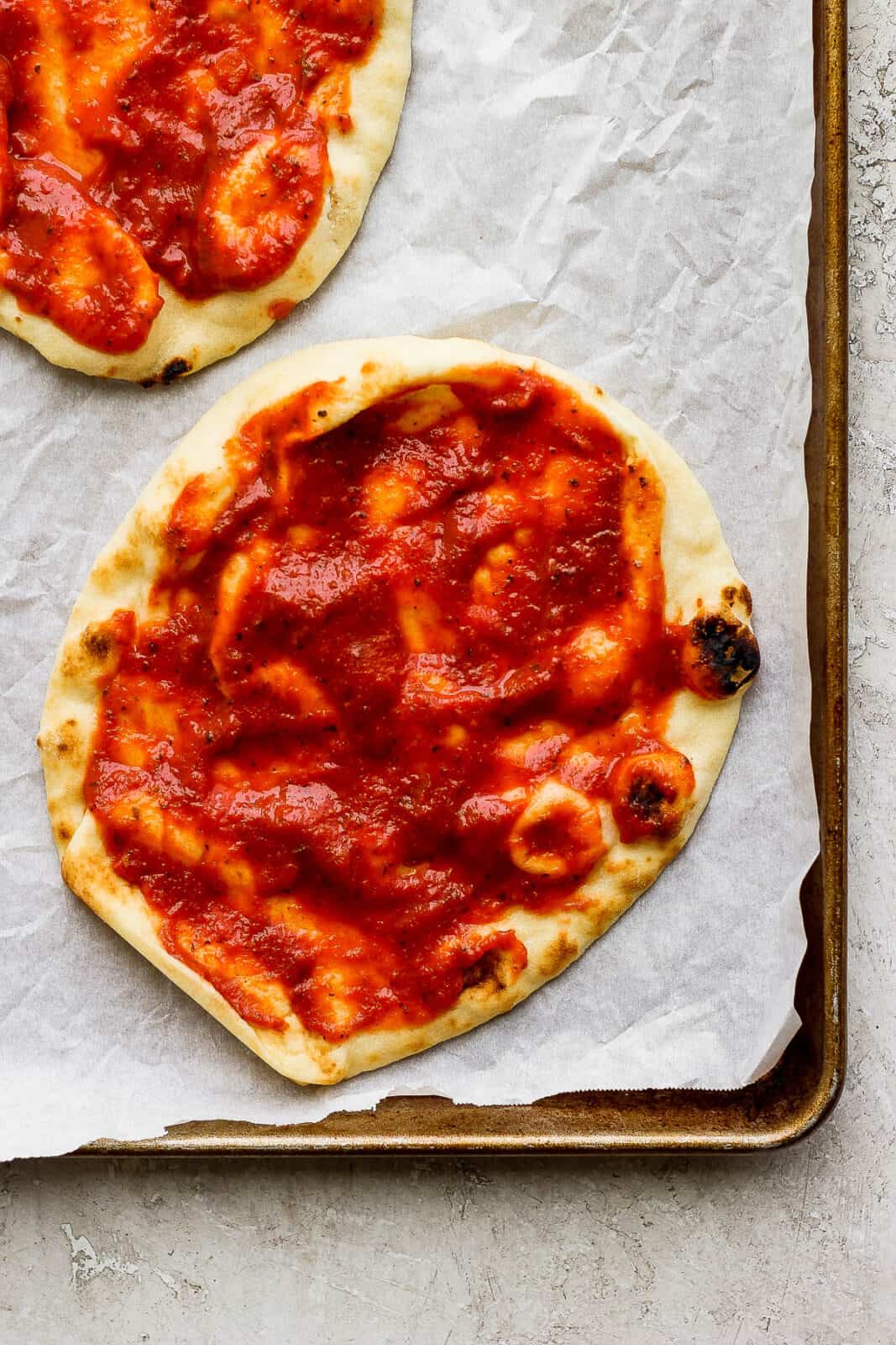 Two pieces of naan flatbread topped with pizza sauce on a baking sheet lined with parchment paper to make naan flatbread pizza.