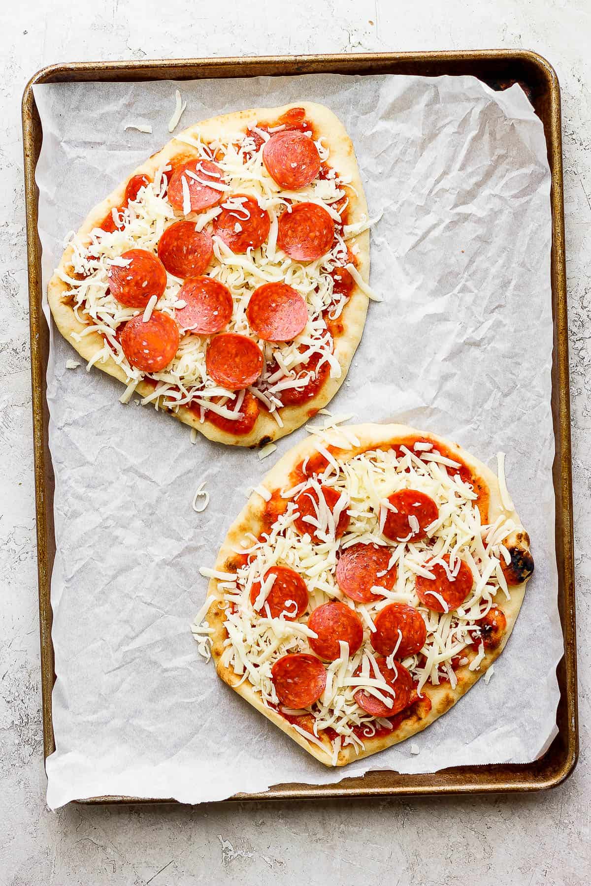 Two naan pizzas on a parchment-lined baking sheet with sauce, cheese, and pepperoni.