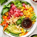 Top down shot of a poke salad in a shallow bowl with tuna, avocado, mango, carrot and a small bowl of sesame ginger dressing.